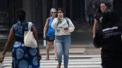 Una mujer vestida con una sudadera blanca y jeans se cubre el rostro con un pañuelo mientras cruza una calle de la ciudad, junto con otros peatones. 