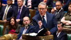 Keir Starmer de traje oscuro habla desde el atril en el Parlamento británico, con otros asistentes sentados en filas de fondo.
