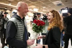 Homem sorridente recebe uma mulher também sorrindo com um ramo de rosas, enquanto diversos câmeras gravam o momento