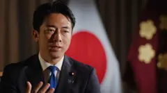 ダークスーツに青いネクタイを着けた小泉進次郎氏が、胸の前で右手を広げ、前方を見て話をしている。後ろには日本の国旗が見える