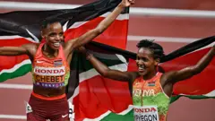 Beatrice Chebet et Faith Kipyegon sourient et traînent des drapeaux kenyans derrière elles tout en célébrant sur une piste de course après avoir remporté respectivement l'or et l'argent au 5 000 m féminin aux Championnats du monde d'athlétisme à Tokyo.