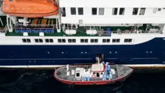 Vista aérea mostra pessoas ajudando pacientes do cruzeiro MV Hondius a entrar em um bote, com o navio ancorado em frente ao porto da Praia, a capital de Cabo Verde, no dia 6 de maio de 2026