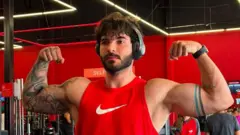 A imagem mostra um homem em uma academia posando com os dois braços flexionados para destacar a musculatura dos bíceps. Ele veste uma regata vermelha com o logotipo da Nike, usa fones de ouvido e um relógio inteligente, além de exibir tatuagens nos dois braços. Ao fundo, aparecem equipamentos de musculação e um ambiente com paredes vermelhas e iluminação em linhas de LED no teto.