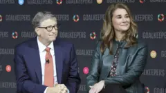 Bill and Melinda Gates seen at an event. They are both looking away but are seated on stools on a stage. 