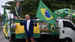 Apoiador do presidente Jair Bolsonaro carregando bandeira do Brasil, em frente a um pequeno caminhão também decorado com bandeiras, bonecos infláveis de Lula vestido de presidiários e uma imagem de Bolsonaro em papelão