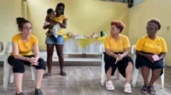 Uma mulher em pé com bebê no colo e outras três mulheres sentadas em roda