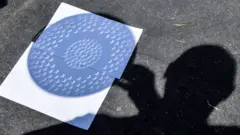 La silhouette d'un homme dans l'ombre tenant une passoire au-dessus d'une feuille de papier blanc, comme autre moyen d'observer une éclipse solaire