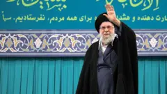 O líder supremo do Irã, Ali Khamenei, usando óculos, barba grisalha e longas vestes escuras, saúda a multidão em Teerã.