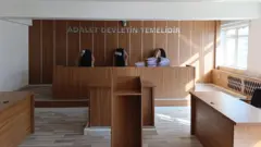Boş bir mahkeme salonunda, mahkeme heyetinin yer aldığı kürsüde dosyaların yığıldığı görülüyor. 