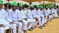 A group of repentant terrorists wearing white garments and yellow camps as dem dey pass out of di rehabilitation programme wey govment organise for dem.