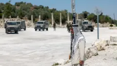 رجل فلسطيني كبير في السن يرتدي سترة بدلة رسمية لونها سكري فوق لباس تقليدي بنفس اللون وعلى رأسه يضع الكوفية (الملونة بالأبيض والأسود)، خلفه مركبات وقوات إسرائيلية وخلفهم تظهر أعمدة أثرية.