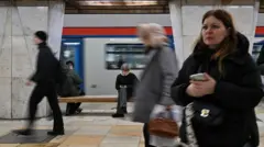 Станция метрополитена. На заднем плане поезд и пассажиры, сидящие на скамейке. На среднем плане пассажиры, идущие справа-налево. На переднем плане справа женщина в темной одежде, с длинными волосами, смотрящая перед собой. Обеими ладонями она держит смартфон.