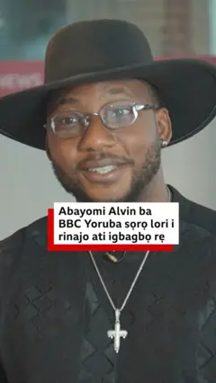Abayomi Alvin wọ asọ shaati dudu, o de fila bẹnti gọ, o si fi ẹgba ọrun sọrun ati igo soju
