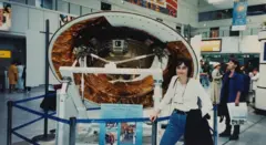 Michele Dougherty, de 30 años, con un top blanco y vaqueros azules, gafas colgando del cuello y una chaqueta negra colgada del brazo. Está de pie delante de una parte redonda de una nave espacial, en parte blanca pero con partes de lámina de cobre. Está delimitada por una barandilla azul. La escena parece transcurrir en un museo, o tal vez en un aeropuerto, y hay gente alrededor.