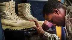 soldado americano cabisbaixo segura em duas botas de combate que estão à sua frente