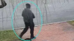 黒っぽい帽子、服を身に着けた容疑者の画像。車道脇らしきところを歩いているように見える。画像では青線で囲まれて強調されている