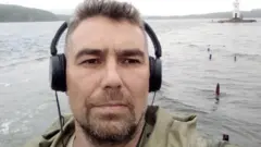 Андрей Акузин в больших черных наушниках фотографирует сам себя на фоне реки и холмов