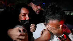 Duas mulheres choram de mãos dadas em meio às operações de resgate após o duplo terremoto na Venezuela, enquanto um homem as abraça por trás.