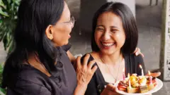 Duas mulheres riem enquanto uma segura um prato com bolos e velas acesas.