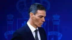 O primeiro-ministro Pedro Sánchez, vestido de terno e gravata, diante de um fundo azul-marinho com o escudo da Espanha