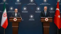 İki erkek kürsülerinin arkasında duruyor. Yanlarında Türkiye ve İran bayrakları var. Arkadaki mavi panoda "Türkiye Cumhuriyeti Dışişleri Bakanlığı" yazıyor. 