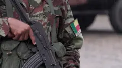 A close-up of a soldier holding an automatic weapon. A badge on the left arm says 'Benin'.