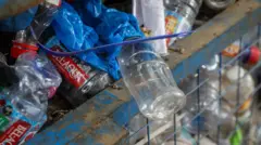 Niebieski metalowy kosz z kratką jest wypchany plastikowymi butelkami w różnych kolorach. Na górze kosza przez krawędź zwisa przezroczysta plastikowa butelka, na której znajduje się niebieski plastik oraz niebieska opaska zaciskowa.