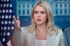 Karoline aponta para a frente enquanto fala em frente a bandeira dos EUA e símbolo da Casa Branca