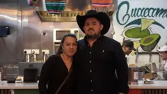 Manolo viste una camisa oscura de botones y un sombrero. Está de pie junto a su esposa, Gabriella, quien viste también una camisa oscura. Manolo tiene un sombrero de baquero. Atrás se ve el mostrador de su negocio con una caja registradora y dos personas con uniforme blanco. La decoración del lugar tiene elementos típicos mejicanos, como sombreros y mantas de colores brillantes, entre naranja, turquesa y amarillo. 