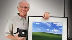 Un sonriente Chuck O'Rear posa sonriente, sujetando la cámara con la que tomó "Bliss" y con una impresión enmarcada de la foto, que muestra una colina verde sobre un cielo azul brillante con nubes blancas y esponjosas.