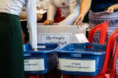 ပလက်စတစ် ပုံးတစ်ပုံး၊ အပြာရောင် ခြင်း ၂ ခြင်းနဲ့ လုပ်ထားတဲ့ မဲပုံးတွေထဲက မဲတွေကို ရေတွက် နေသူတွေ 