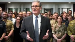 Sir Keir Starmer ahagaze imbere, yambaye ikositimu na karuvati n'indorerwamo, akoze ikimenyetso n'ibiganza, akikijwe inyuma no ku mpande n'abo mu nzego z'umutekano barimo n'abambaye imyenda ya gisirikare. 