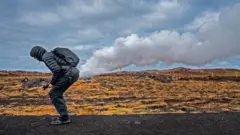 Kısa şişme montlu, kapüşonlu, yürüyüş pantolonu ve ayakkabı giymiş bir adamı gösteren arşiv fotoğraf. Öne doğru çömelmiş ve sanki poposundan bir bulut patlaması geliyormuş gibi konumlanmış. Çimenli bir arazinin yanındaki bir yolda.