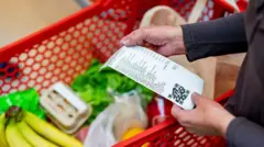 Pessoa segura nota fiscal em frente a carrinho de mercado com produtos alimentícios