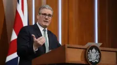 İngiltere Başbakanı Starmer bir kürsüde konuşurken elini kaldırmış. Üzerinde siyah bir takım elbise, beyaz bir gömlek ve yeşil bir kravat var. Arkasında Birleşik Krallık bayrağı görülüyor