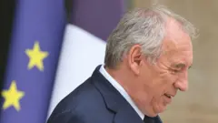 Франсуа Байру, седой пожилой мужчина, стоит, склонив голову, на фоне флага ЕС 