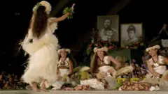 Dançarinas de hula se apresentam ao público frente a dois retratos reais