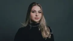 Raphaela Coiado é loira e usa blusa preta e colar prata. Ela olha para a câmera sem sorrir.