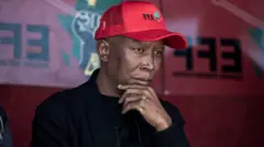 Julius Malema wear red cap and black shirt. E put im hand for im chin.