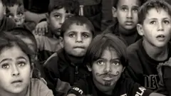 صورة بالأبيض والأسود لأطفال صغار يجلسون على الأرض وينظرون إلى الأعلى، وقد رُسم على وجه أحدهم شارب ولحية بالقلم.