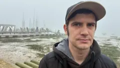 Justin Francis é um jovem vestindo um casaco escuro com capuz e um boné de beisebol. Atrás dele, vê-se o lodo e os barcos do porto de Brightlingsea em um dia de neblina.