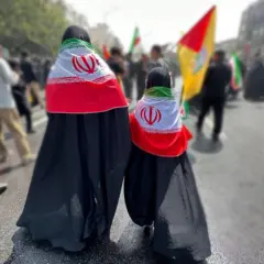 فتاتان تسيران إلى الأمام ويظهر ظهرهما، و ترتديان عباءة سوداء وتغطيان ظهرهما بعلم إيران.