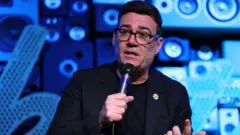 青い背景の檀上でマイクを持って話すバーナム氏。黒縁のめがねをかけ、黒いジャケット、黒い丸襟のトップスを着ている