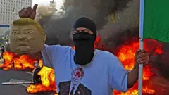 Manifestante com balaclava segura máscara de Donald Trump em frente a veículos incendiados na rua