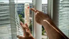 Дві руки, які відхиляють жалюзі на вікні і показують на термометр, прикріплений до рами. На шкалі видно високі значення — приблизно близько +50 градусів. За вікном — багатоповерхові житлові будинки і зелень у дворі.