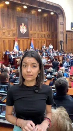 Novinarka gleda u kameru, iza nje sednica skupštine.