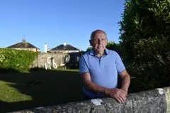 Un hombre con un polo azul junto a un muro de piedra que le llega a la cintura. Al fondo, una franja de césped se extiende hasta un alto muro perimetral. Parte del muro está cubierto de hiedra; en otra parte se puede ver el número 796 en grandes letras blancas de madera. El hombre en primer plano es mayor, calvo y tiene las manos apoyadas en el muro bajo. Es de día y el cielo es azul claro.