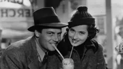 Siyah beyaz eski bir fotoğrafta bir çift, ağızlarına götürdükleri pipetle bir bardaktan içecek paylaşıyor