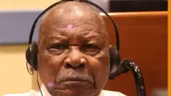 Félicien Kabuga areba muri 'camera' yambaye inyumvisho (écouteurs) mu matwo. Yambaye ishati y'umweru, afite umusatsi mucye w'imvi n'ubwanwa bucye bwo hejuru bw'imvi. 