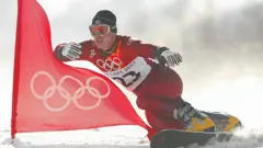 Ryan Wedding en una snowboard con el uniforme rojo de los Juegos Olímpicos de Salt Lake City, gafas de nieve, botas amarillas y un gorro negro. A su lado ondea una bandera roja con el logotipo olímpico en blanco.